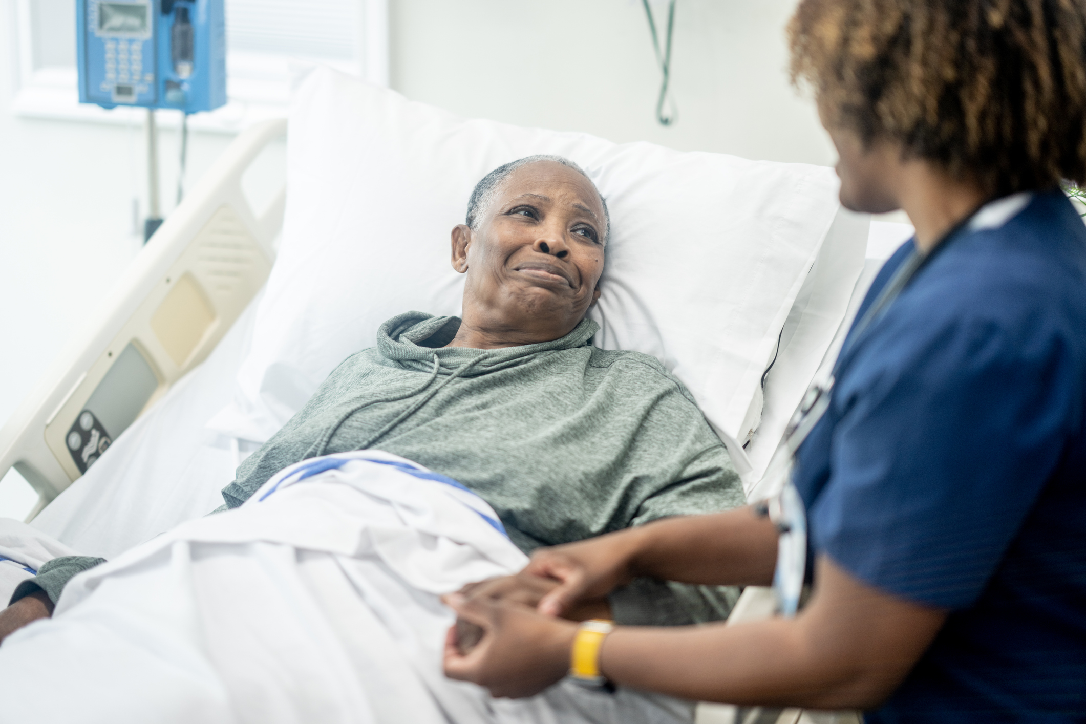 A female nurse of African decent checks in on her senior patient as she awaits to be called for surgery. She is smiling as she reassures her patient and puts her at ease before the procedure.